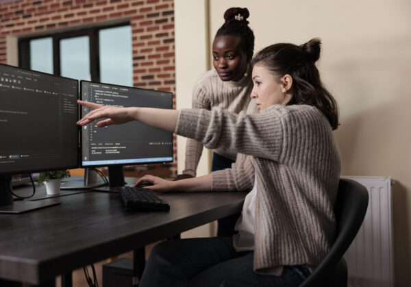 Caucasian network developer pointing out high usage of processing power in mainframe grid. African american programmer helping coworker to solve overloaded system storage space.