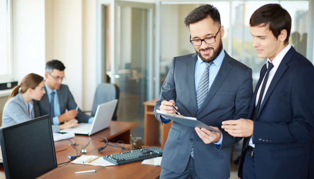 Confident businessman with clipboard consulting his partner on background of their colleagues in office