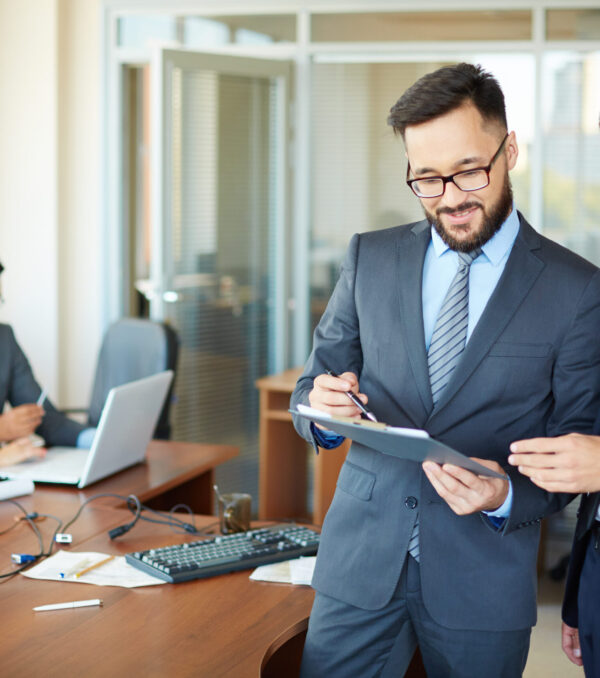 Confident businessman with clipboard consulting his partner on background of their colleagues in office